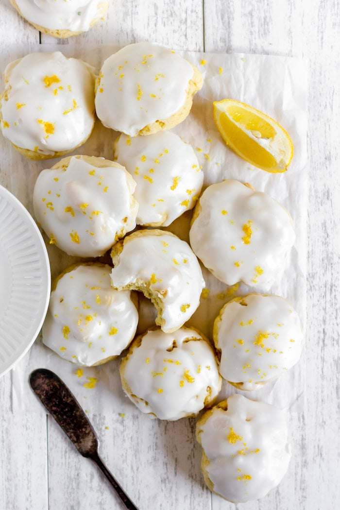 Photo, taken from above, shows about 10 iced Italian lemon cookies on a parchment paper on a table. A wedge of lemon sits nearby.