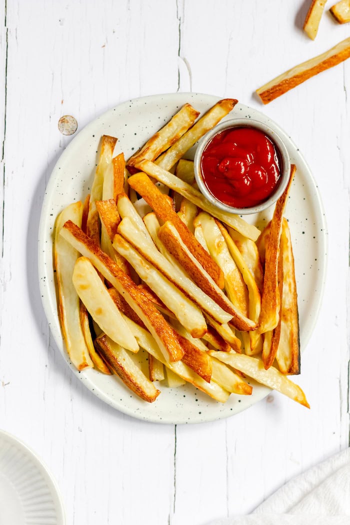 Image, taken from above, shows a white plate on a white table with oven fries piled on it and a small ramekin of ketchup next to the fries.