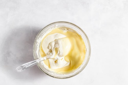 Image, taken from above, shows a glass bowl with whipped cream, with condensed milk and peach flavoring being folded in.
