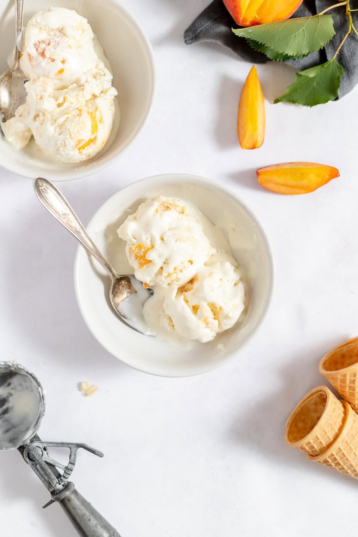 Image, taken from above, shows two white bowls, each with a serving of peach cobbler ice cream in it. Next to the bowls sits a few slices of peaches.
