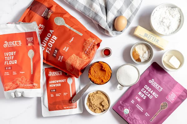 Image shows the ingredients for gluten free pumpkin cookies- quinoa flour, teff flour, arrowroot powder, cacao nibs, butter, egg, flour, sugar, pumpkin, brown sugar, and more. 