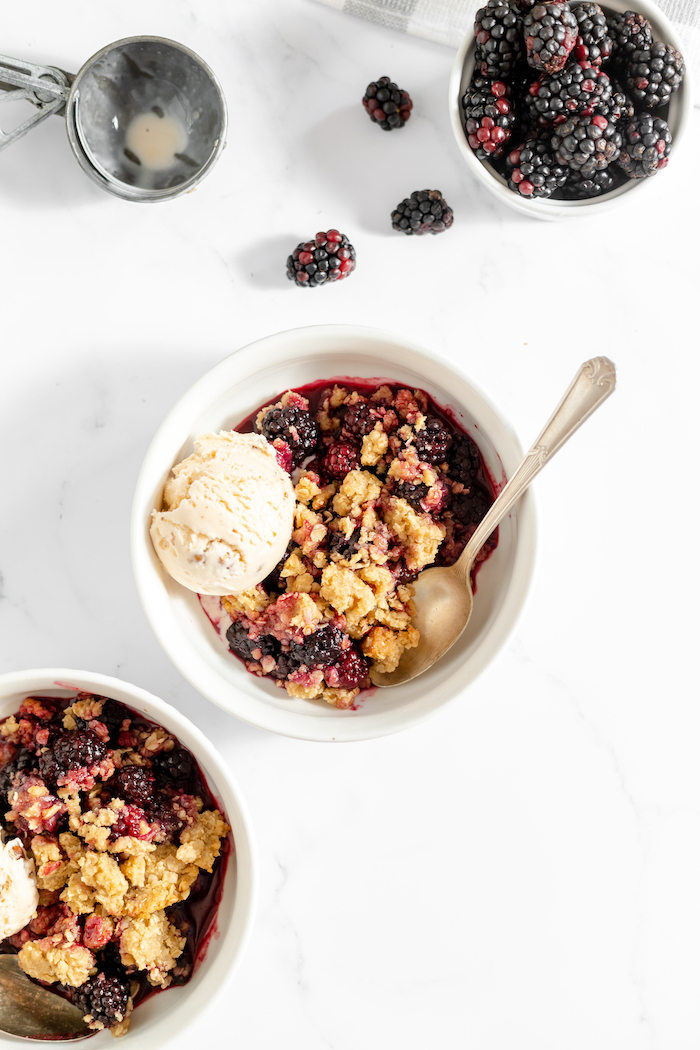 Photo, taken from above, shows several bowls on a counter. In two are blackberry crisp with a scoop of vanilla ice cream. The third holds large blackberries.