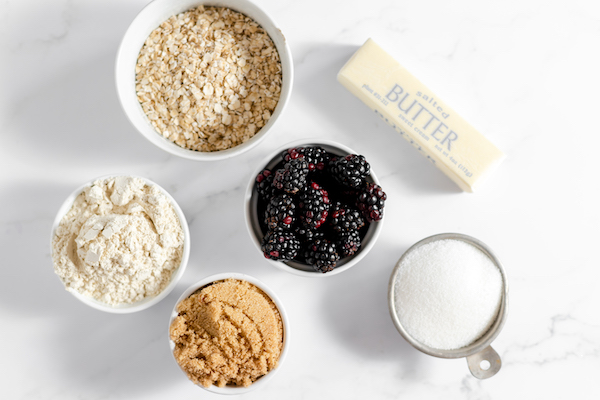 Image shows several bowls on a counter full of the ingredients to make gluten free blackberry crisp, including oatmeal, brown sugar, sugar, butter, oat flour and blackberries. 