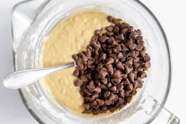 Image, taken from above, shows a glass mixing bowl with chocolate chips added to the muffin batter, with a spoon to mix them in.