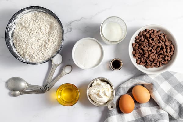 Image, taken from above, shows a countertop covered in the ingredients needed to make gluten free chocolate chip muffins. A bowl of flour, some salt, sugar, eggs, oil, chocolate chips and more. 