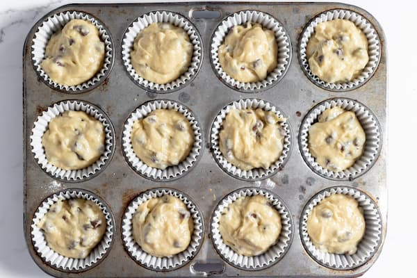 Image shows a 12-cup muffin tin with the chocolate chip muffin batter divided into each cup.