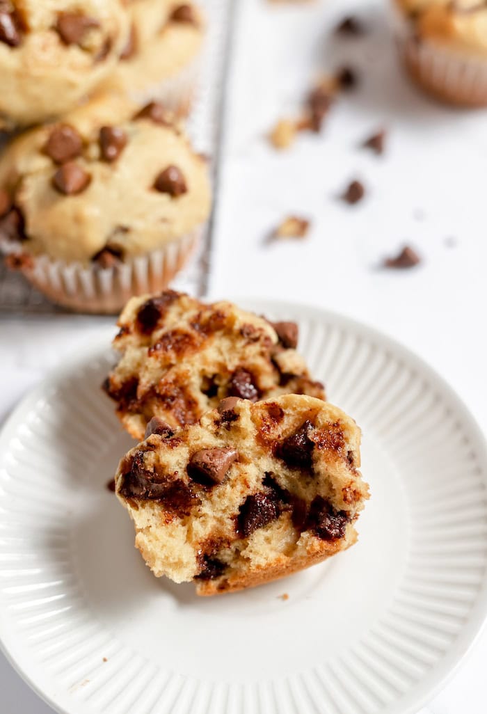 Image shows a chocolate chip muffin torn in half sitting on a small white plate. In the background is a cooling rack with more muffins, and some chocolate chips scattered on the countertop