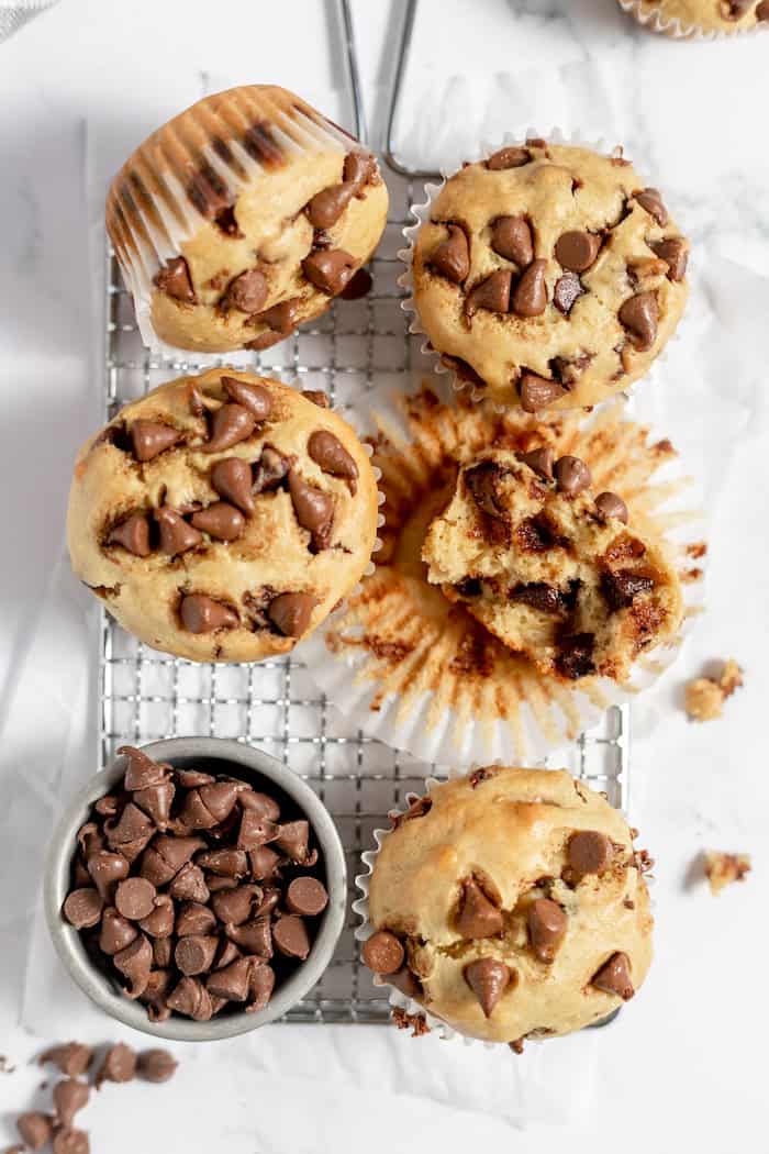 Image shows a close up of several gluten free chocolate chip muffins cooling on a cooling rack. One is split in half showing some melting chocolate chips.