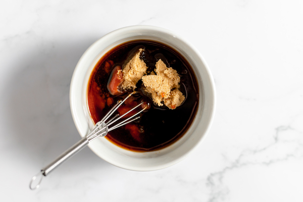 Image shows a white bowl sitting on a counter with soy sauce, onion salt, and Sriracha to make bacon jerky marinade.