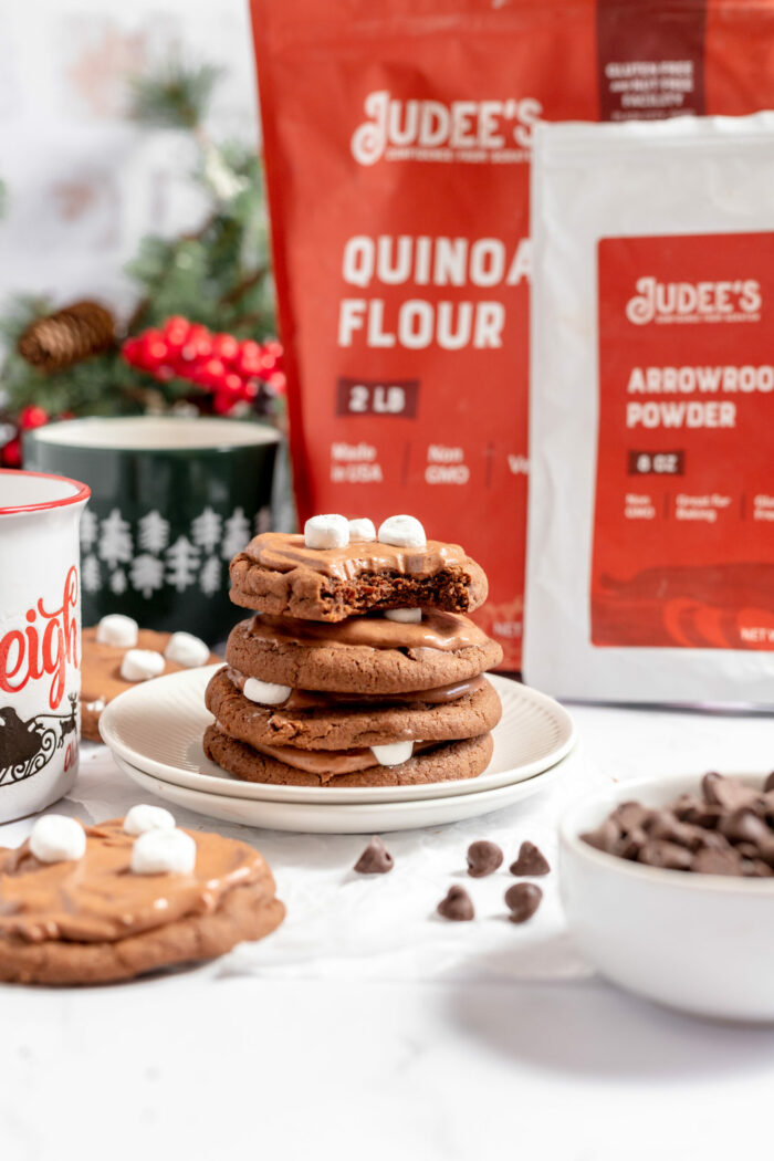 Image shows a pile of gluten free hot chocolate cookies on a white plate with Judee's gluten free products behind it. Chocolate chips are scattered nearby on the counter. 