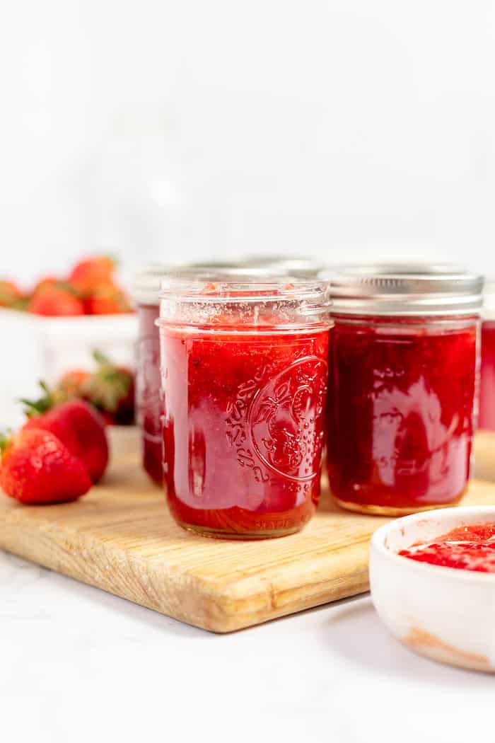 Image shows several jars of strawberry jam on a wooden cutting board. Fresh strawberries sit nearby on the board. 
