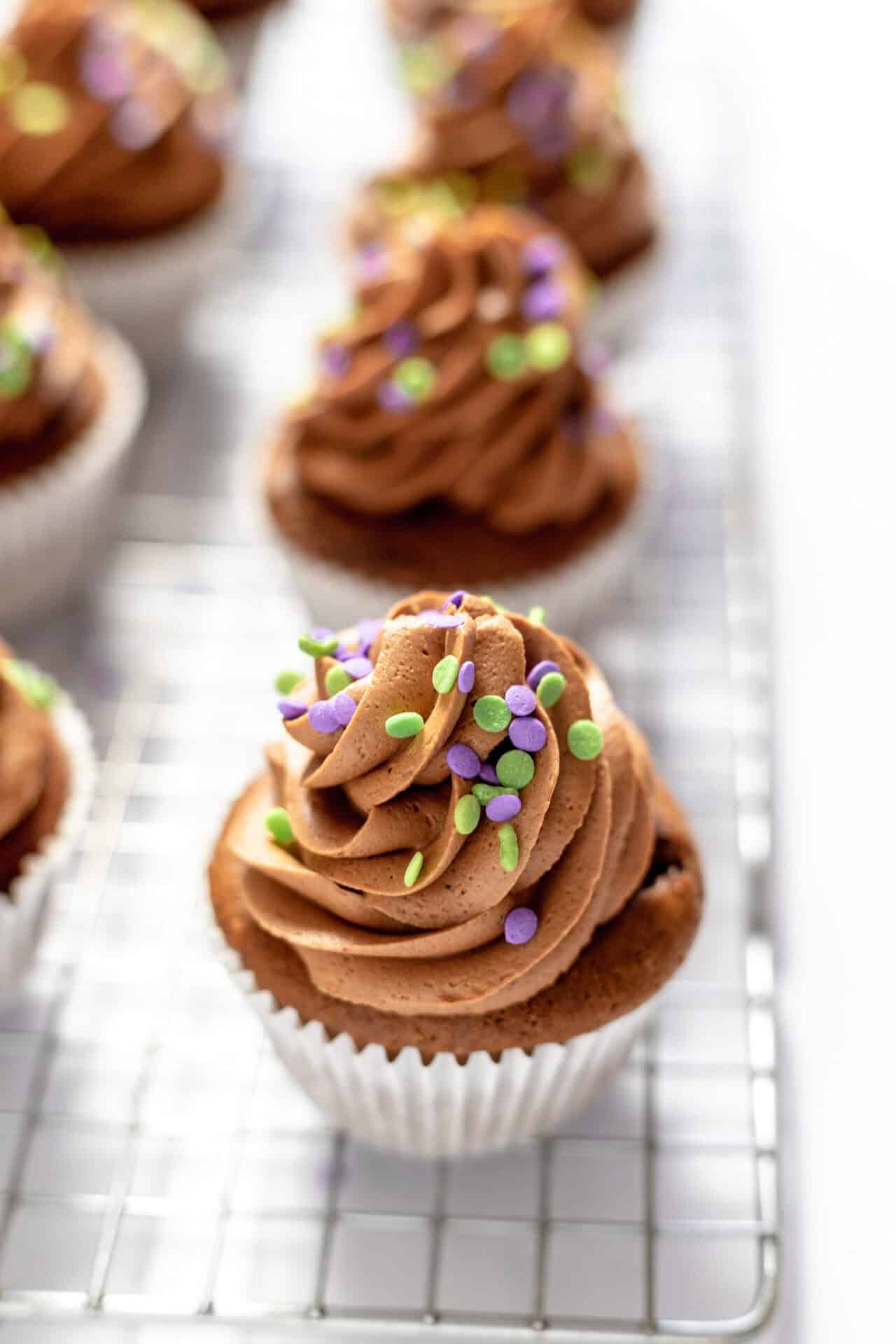Image shows a close up of a gluten free chocolate cupcake topped with chocolate buttercream icing and sprinkles. Several more iced cupcakes sit nearby on the wire rack.