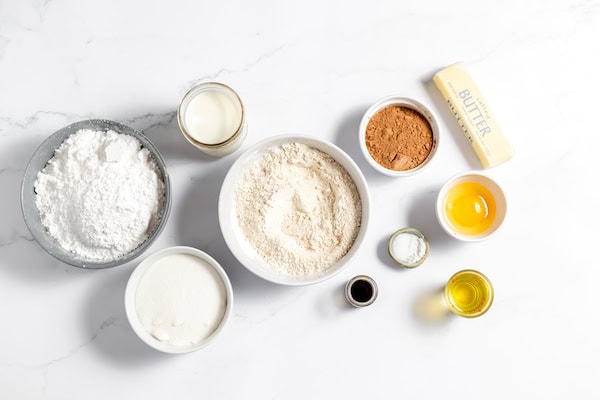 Image, taken from above, shows several bowls full of the ingredients needed to make gluten free chocolate cupcakes, sitting on a white table. Includes several bowls of gluten free flours, sugars, cocoa, egg, oil, butter, and more.