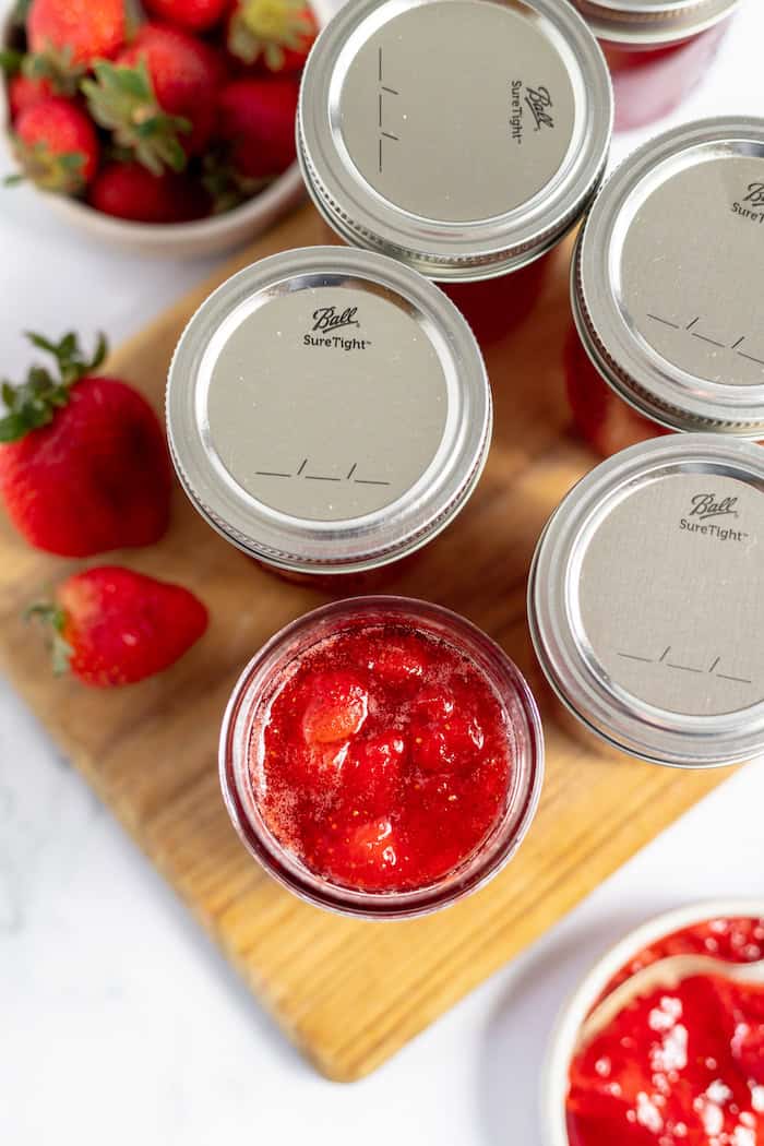 Photo, taken from above, shows the tops of several mason jars of strawberry jam. A small bowl of fresh strawberries sits nearby on the table. 