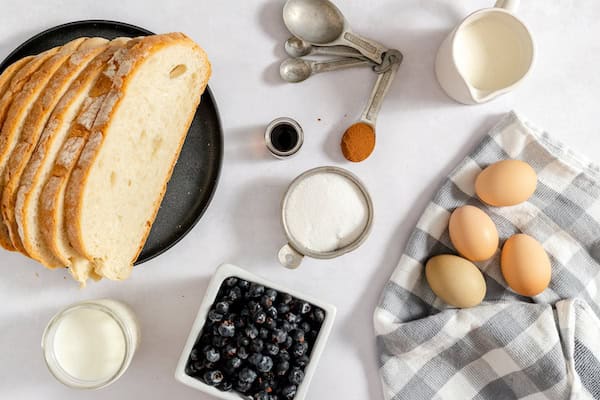 Image, taken from above, shows the ingredients needed for the french toast casserole - gluten free bread, blueberries, milk, sugar, eggs, and spices sitting on a table.