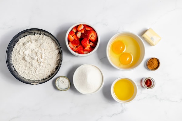 Image, taken from above, shows several bowls full of the ingredients to make gluten free strawberry muffins- flour, sugar, eggs, oil, strawberries, butter and seasonings.
