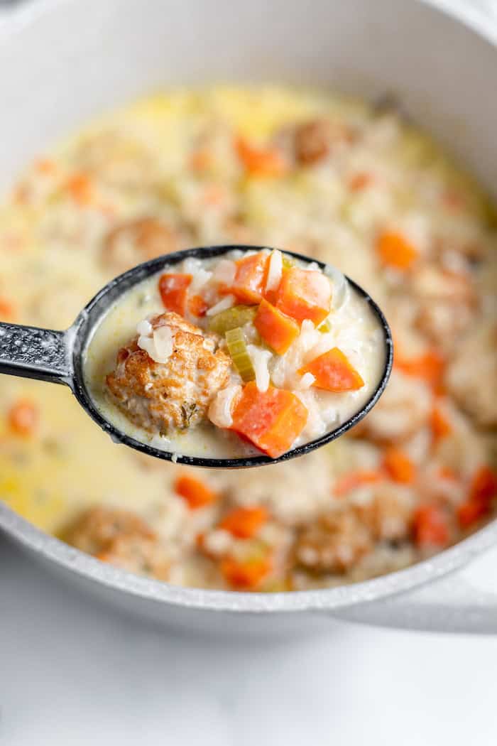 Image shows a close up of a ladle of soup hovering over a pot of chicken meatball soup. 