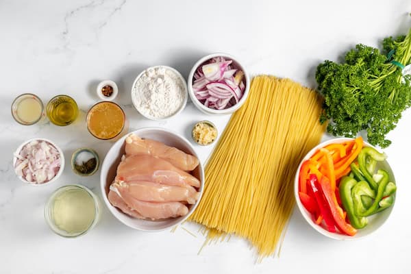 Image, taken from above, shows the ingredients for making chicken scampi laid out on a white counter top. 
