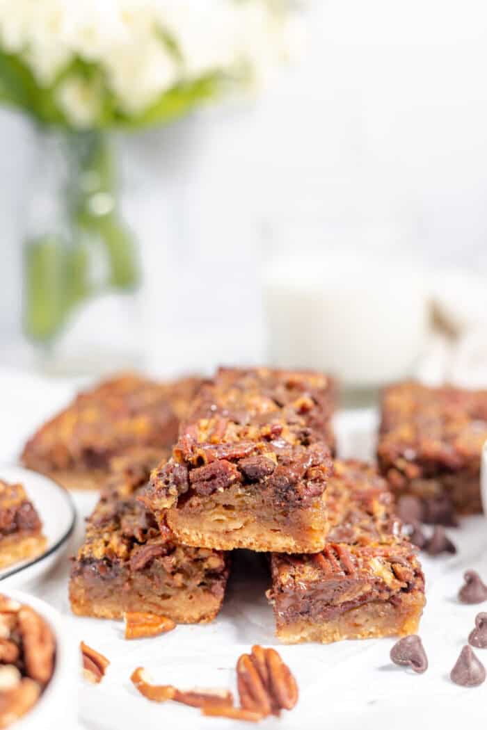 Image shows a stack of gluten free chocolate pecan pie bars stacked on a white plate. Some pecans and chocolate chips are scattered nearby. 