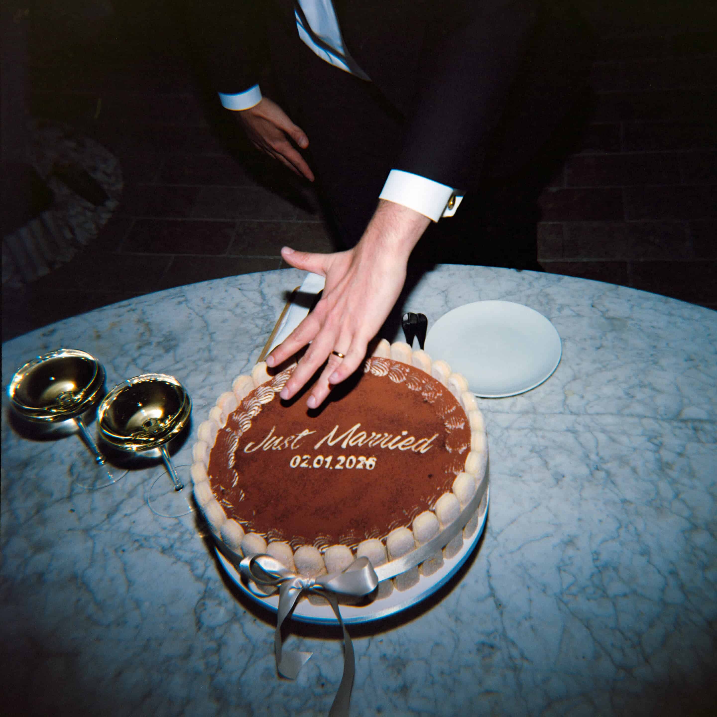 Wedding cake with 'Just Married' and date, hand placing cake topper, wedding rings nearby.