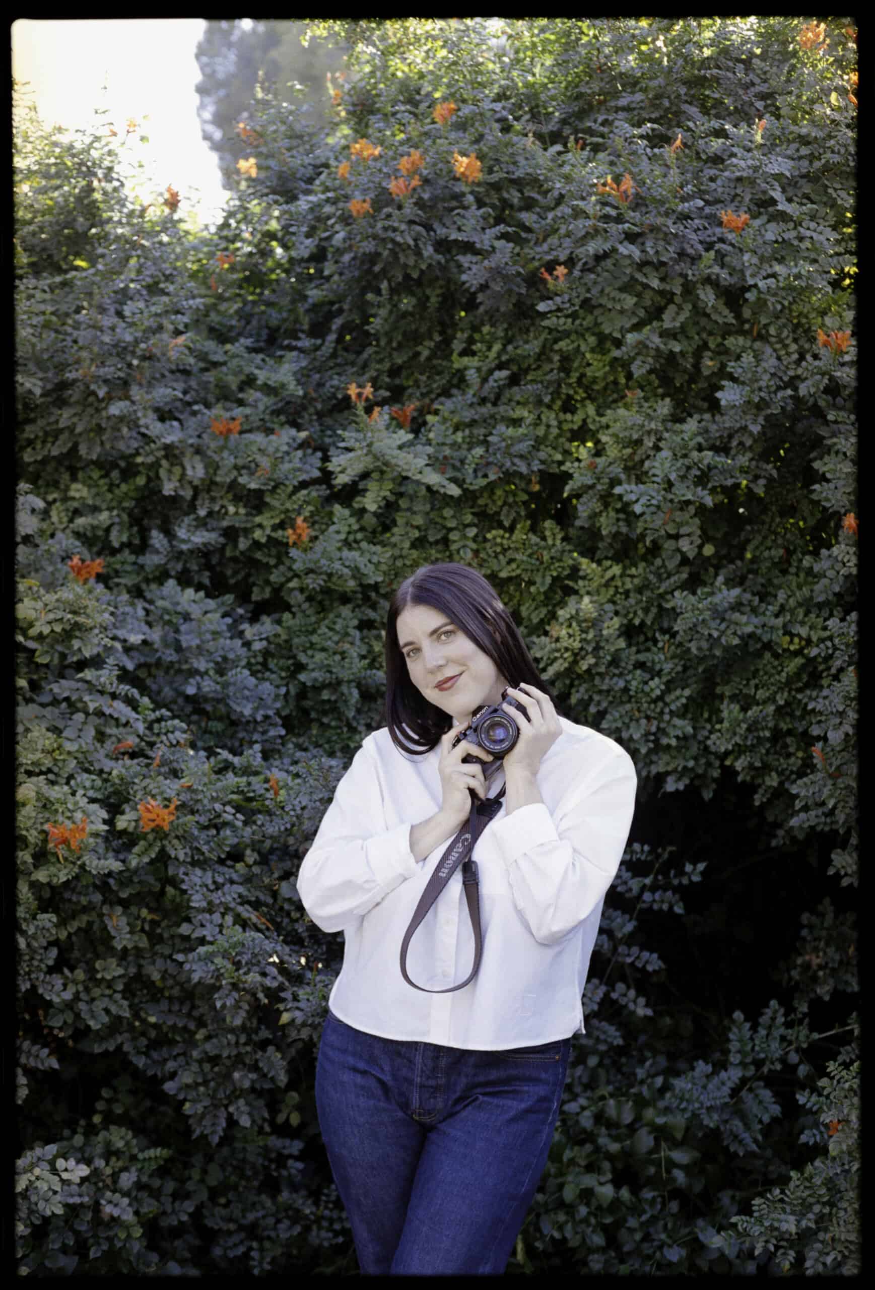 Portrait of a woman holding a camera outdoors with lush greenery and orange flowers.