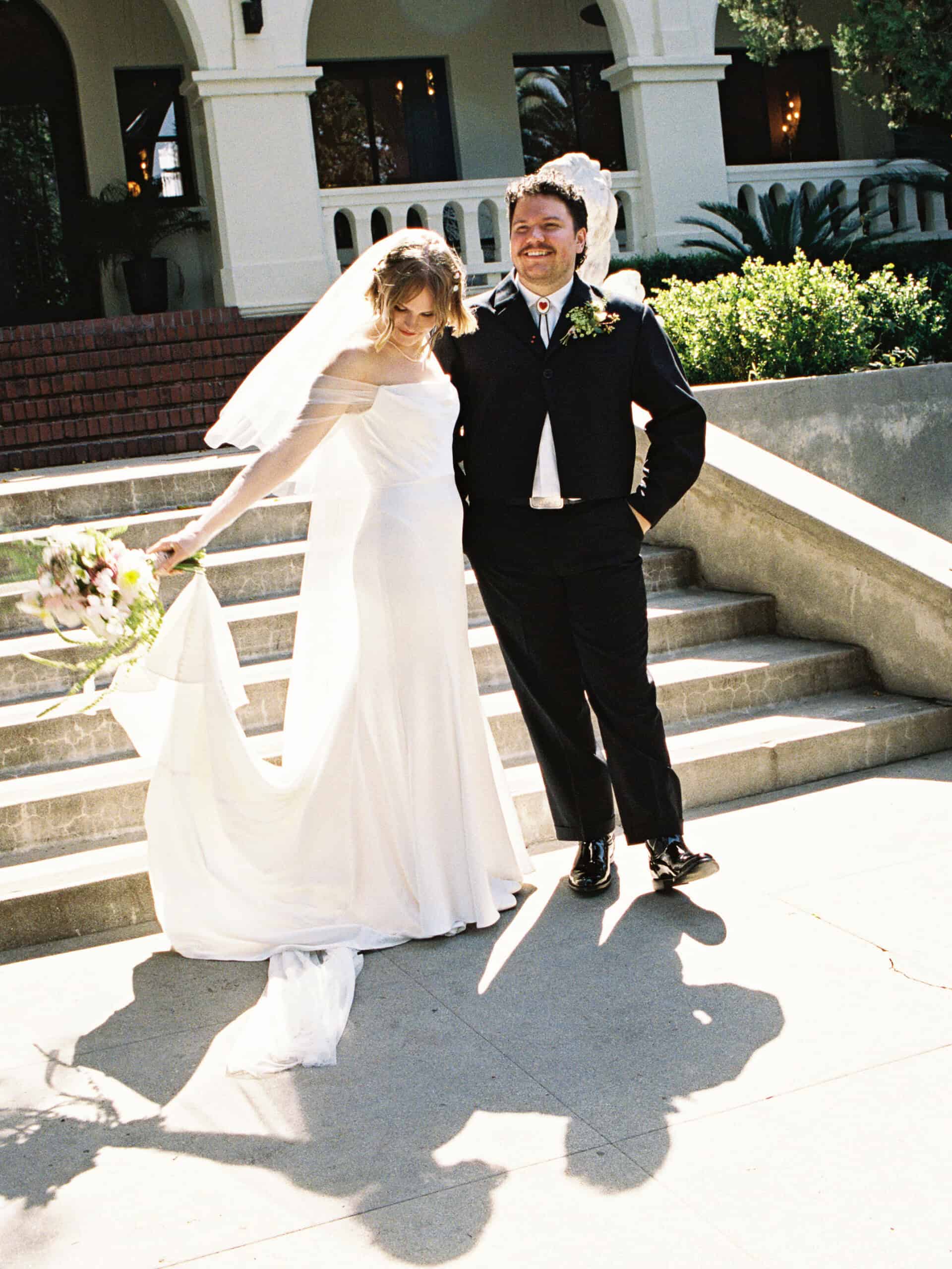 Bride and groom smiling outside on wedding day with sunlight and greenery.