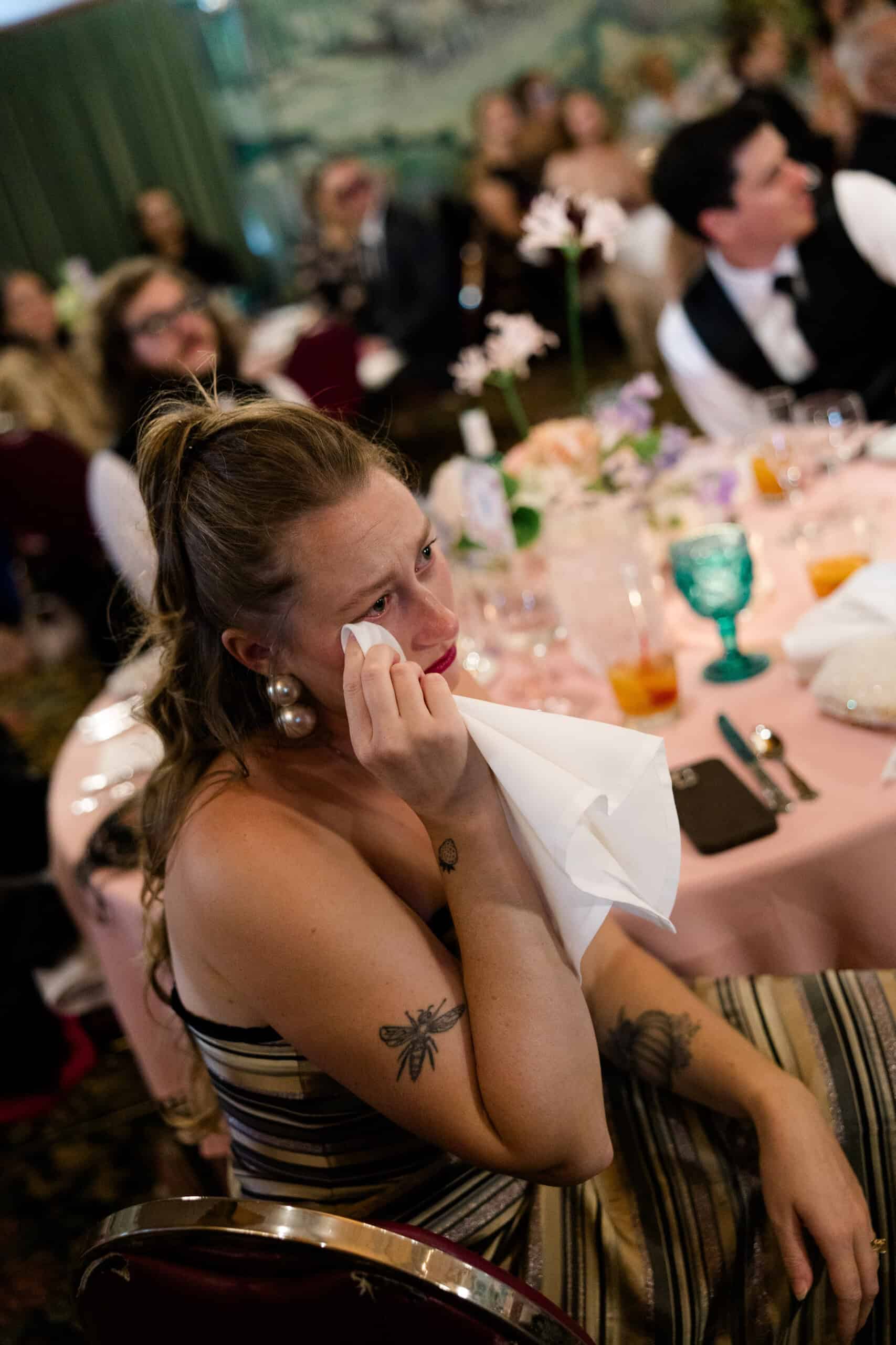 Emotional woman wiping tears at a formal event with guests in the background.