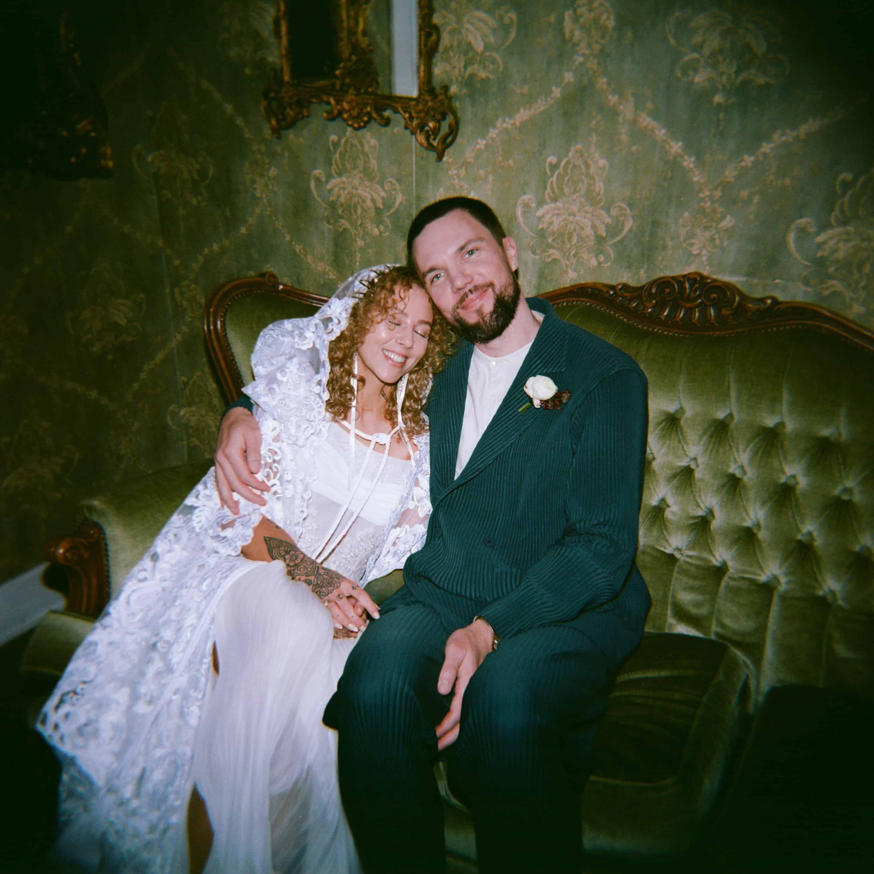 Bride and groom sitting on vintage green sofa in wedding attire.