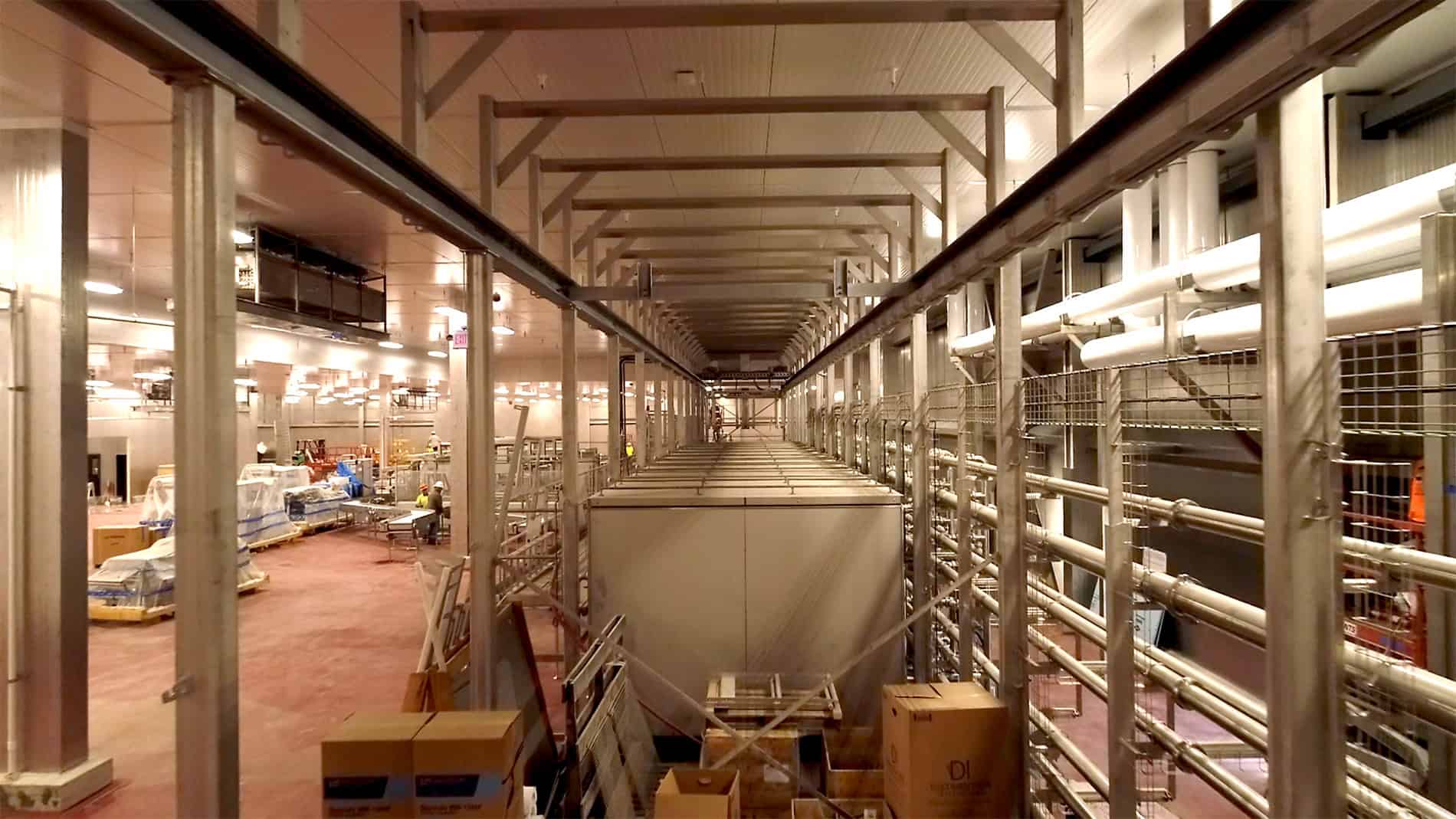 A view from between a long metal grid surrounding square equipment used by the Ruprecht Meat Company Processing Facility. On the left is more equipment and employees in hard hats with pallets of supplies covered in plastic.