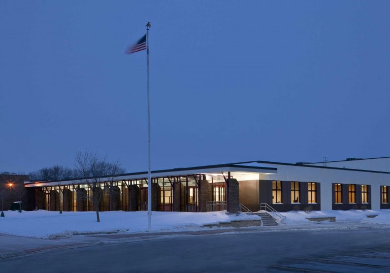 Montini Catholic High School’s exterior at night casts a warm light on the snow outside from the many large windows on the one-story part of the building. A flag pole stands tall in the foreground. Keeley Construction was instrumental in the school’s renovation and upgrade.