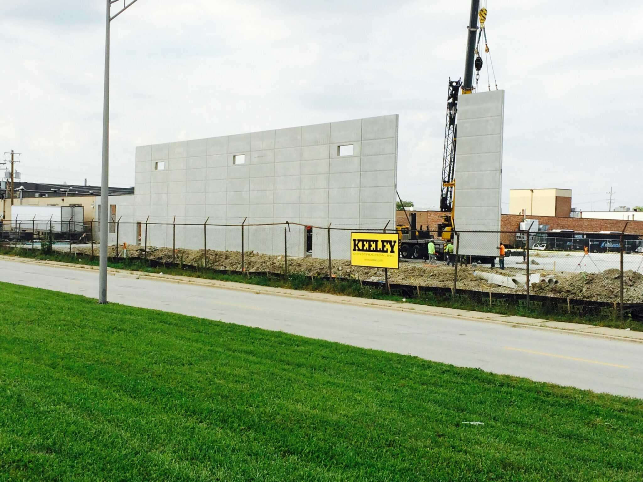 A yellow sign reading “Keeley” is posted on a chain link fence running along a street. Behind the fence, Keeley Construction employees install a tall concrete facade.