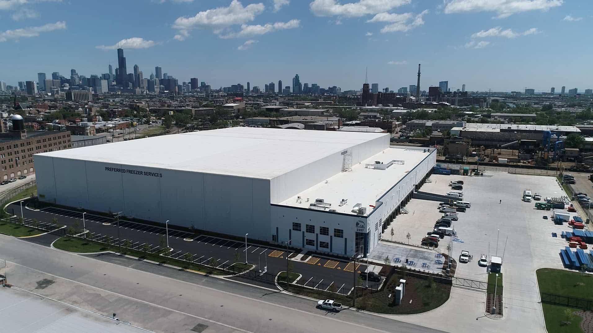 An aerial view of an angled corner and white side of the third Preferred Freezer Services facility built by Keeley Construction Inc. as it dominates an industrial area near a major city visible in the background.