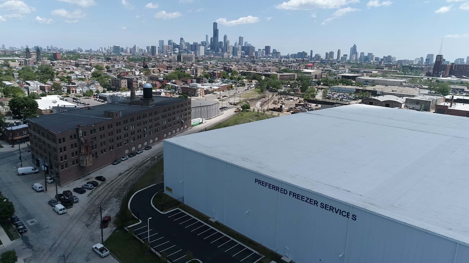The windowless, white side of a large industrial building lies in stark contrast to the old brown and smaller facility across from it. The words on the site of this new warehouse say, “Preferred Freezer Services.” Both buildings are part of business and residential areas seen in the background.