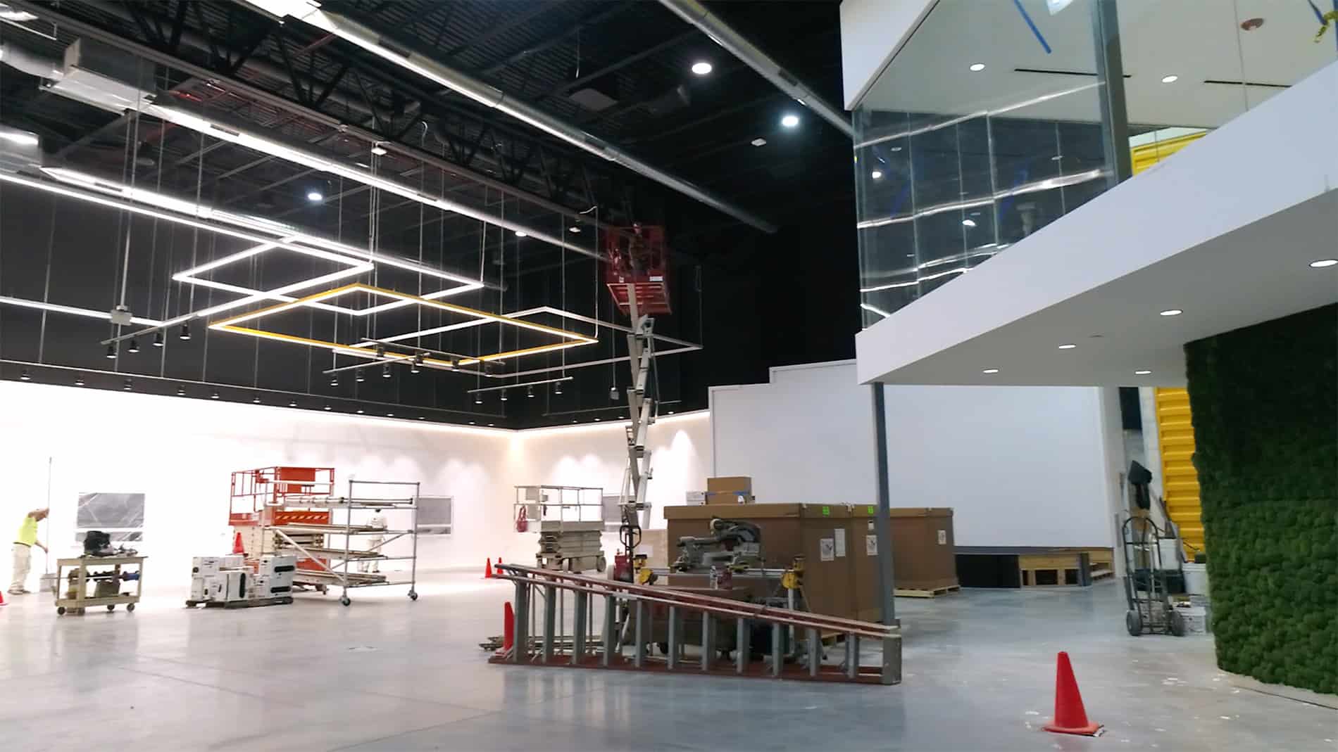 A large room with a large white wall and a black ceiling. Ladders and construction equipment are in the foreground and background.