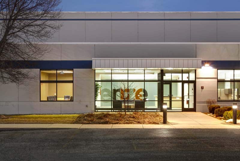 “Lenze,” written on an interior frosted glass wall, is visible from the warm exterior of the Lenze building at night. The entrance to the corporation is inviting and full of windows. A tree stands to the left of the welcome area.