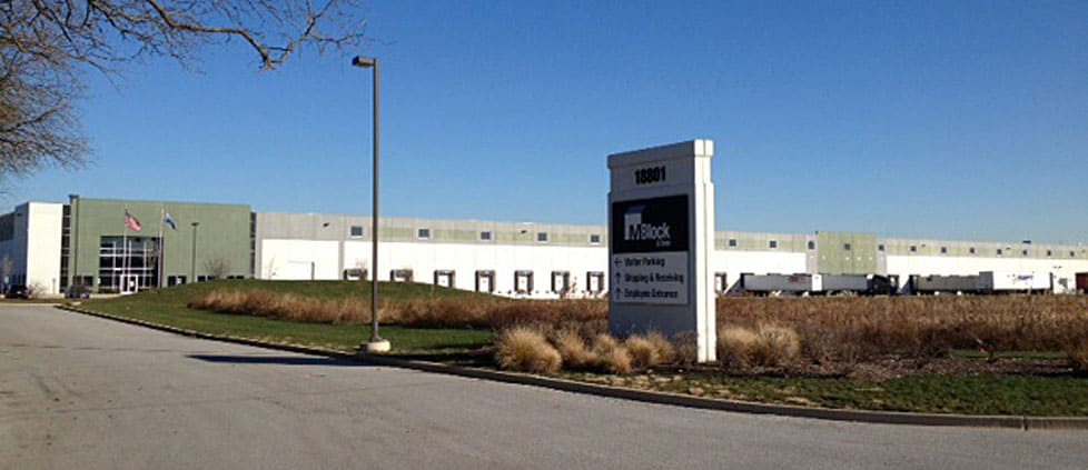 A view from the entrance road to the M. Block and Sons’ corporate building built by Keeley Construction, Inc. shows a tall directional sign that reads “M. Block and Sons” with arrows pointing to different areas of the development, like “visitor parking” and “shipping and receiving.” The parking lot and the large corporate building are in the background.