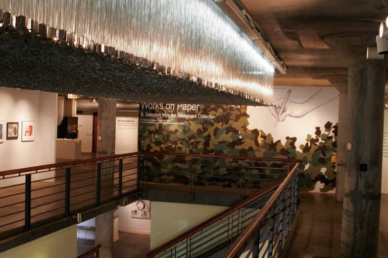The second floor of the National Vietnam Veterans Art Museum has a camouflage mural on the far wall that reads “Works On Paper: A Selection From The Permanent Collection.” Art exhibits line the other walls and the back room, and a prominent, mossy, rectangular light fixture hangs above the open stairwell.