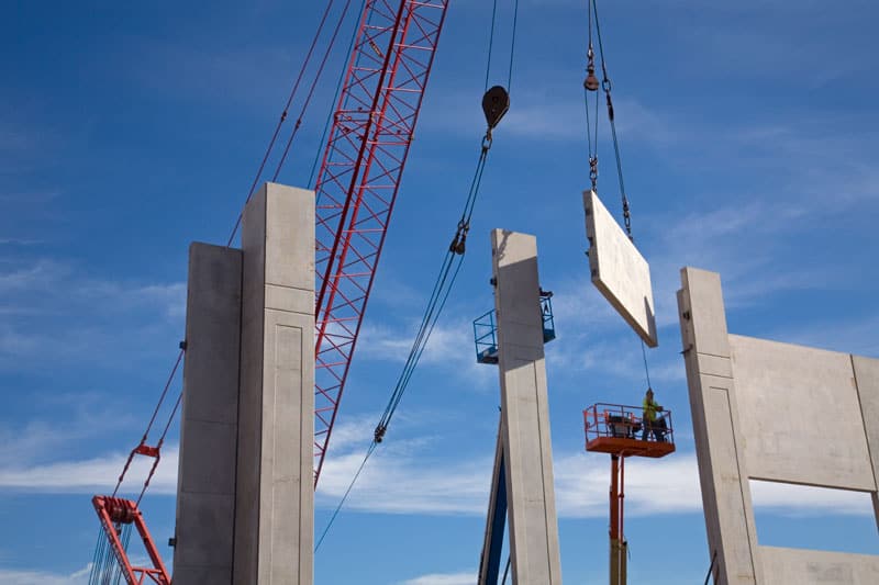 A Keeley Construction crane lifts a concrete wall high in the air to be placed in the wall structure being built. A worker stands on a lift behind the partially built facade for Prologis, a company in the O’Hare Industrial Park.