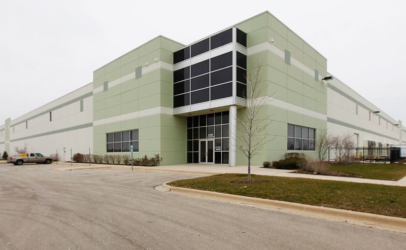 The corner of Wilton Industries' 500,000 square foot warehouse showcases a large entrance full of dark windows. The driveway to the facility is in the foreground