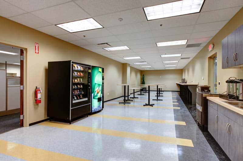 A long break room in the Zone's building is clean and includes tables, chairs, and two vending machines against the wall.