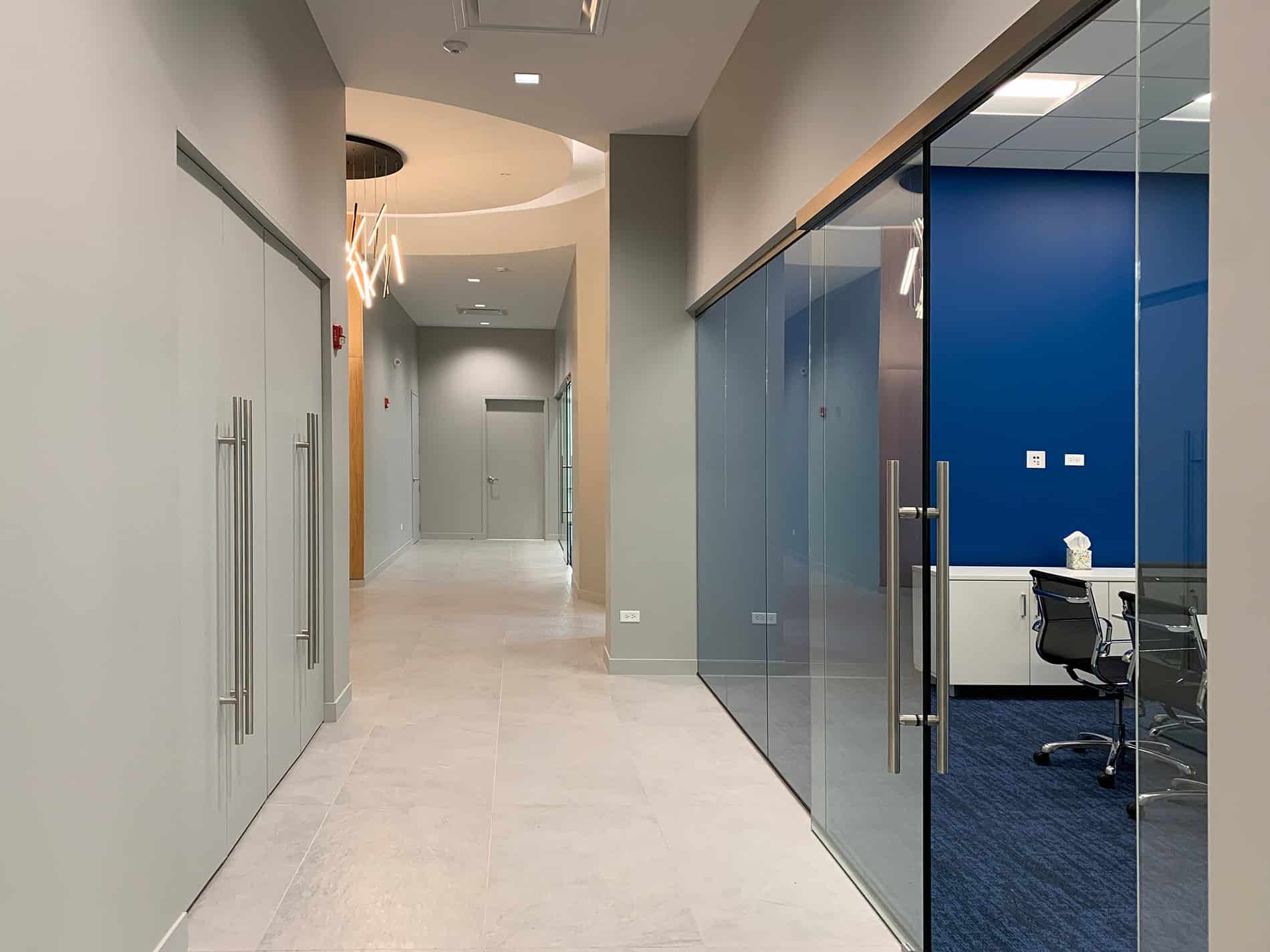 A long hallway with a blue wall and chairs in a room to the right.