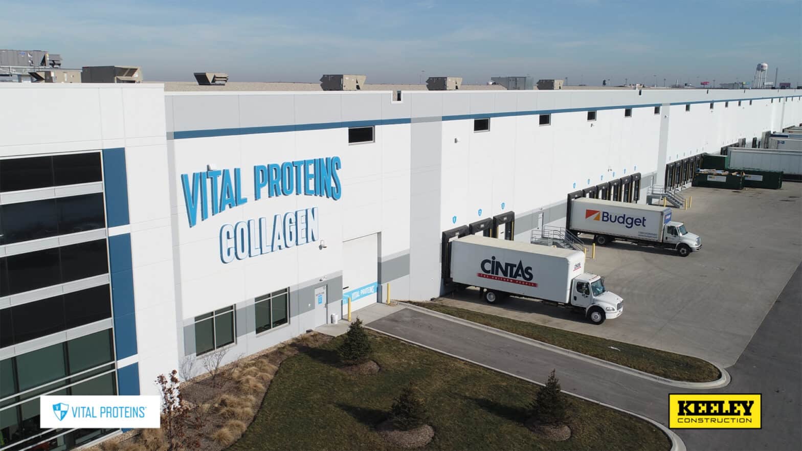 An aerial view of the outside of a long distribution area with docks and semi trucks. A large sign on the side says, "Vital Proteins Collagen."