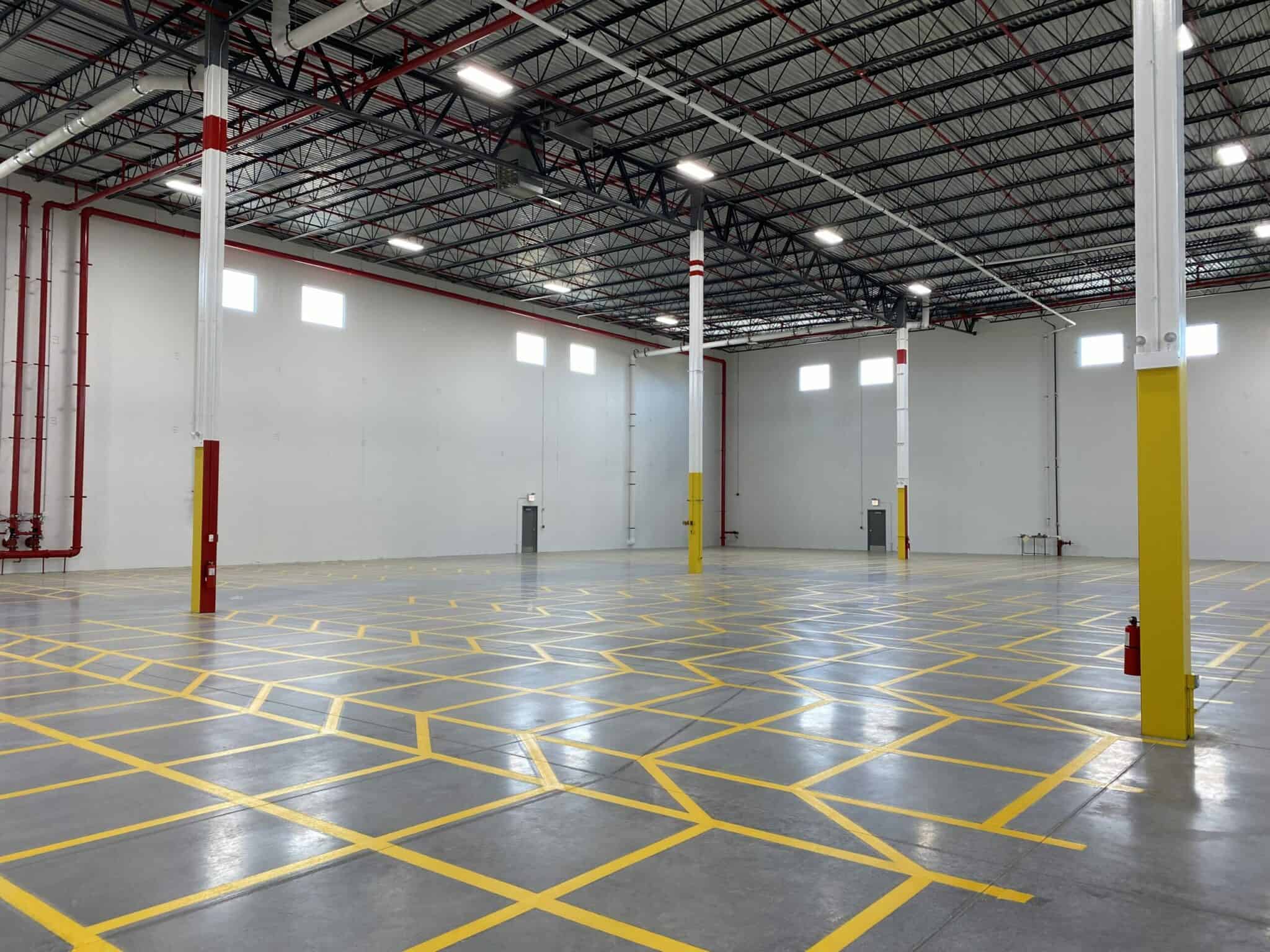 Yellow lines on the floor of the new warehouse in the Nalco Chemical building surround structural poles, red pipes and gray doors on the tall white walls of the large empty space.