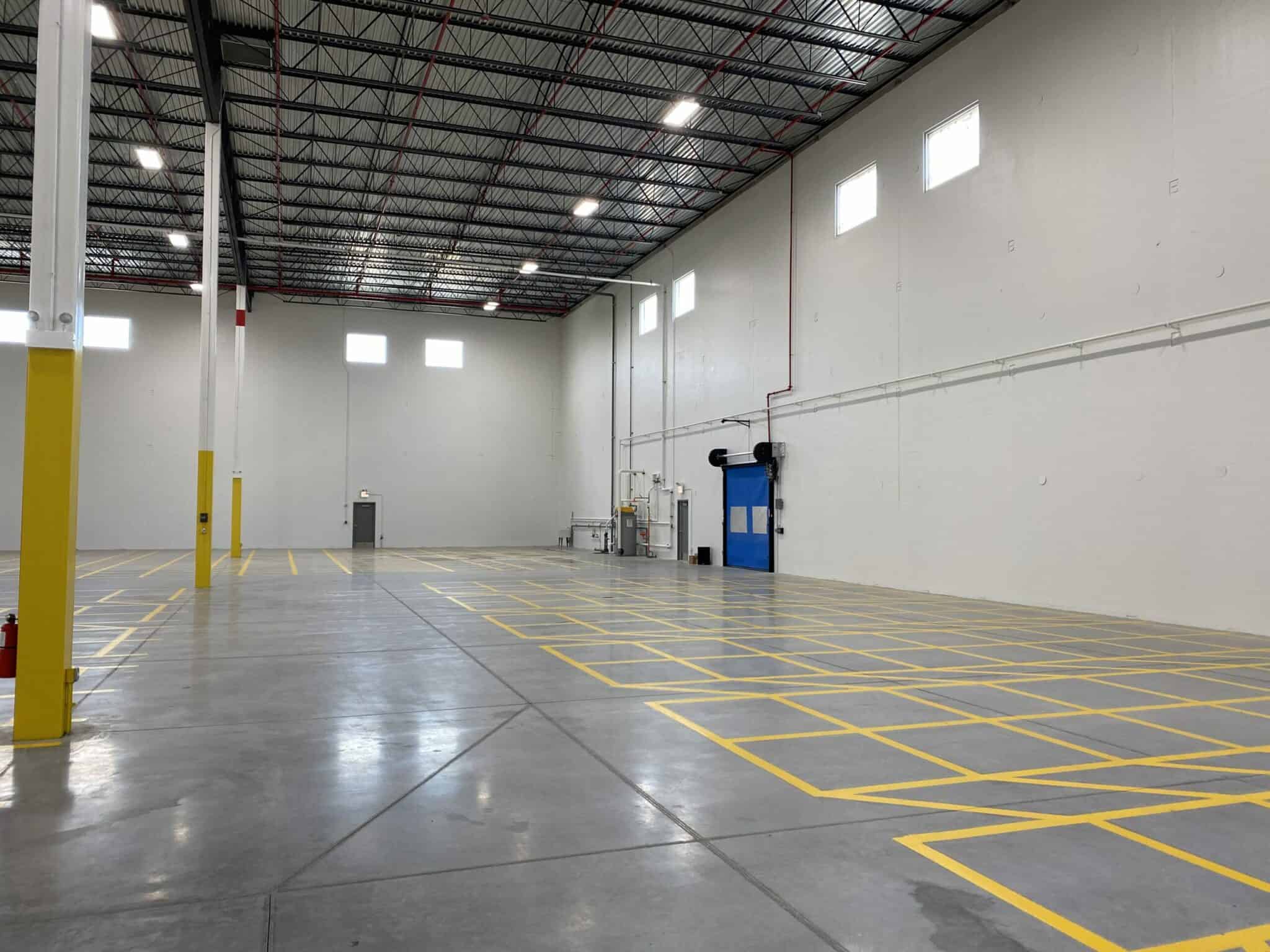 An empty, new warehouse space in the Nalco Chemical building built by Keeley Construction. A closed blue bay door is in the side wall among yellow floor markings in the brightly lit shipping and delivery area.