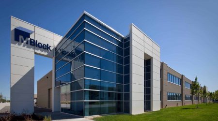 M. Block and Sons' large, new corporate building has floors of windows mirroring the blue sky. A stand-out structure has the words "M. Block" on the top with a sidewalk underneath it. The rest of the building has walls made of brick and other materials that designate the division between corporate offices and warehouse areas.