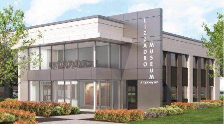 Behind a manicured walkway lined with small bushes sits a modern gray building with clean lines and meeting rooms built with floor-to-ceiling windows and a vertical sign that reads “Lizzadro Museum of Lapidary Art.”