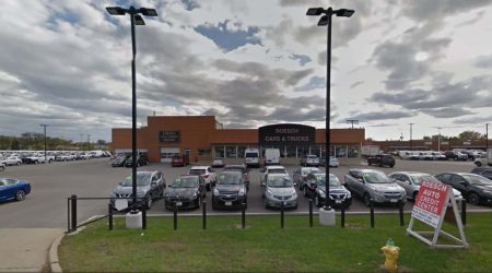 The parking lot and entrance to an auto dealership's showroom are behind a grass verge and two tall street lights.