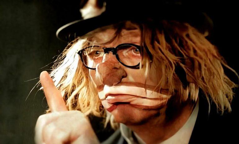 Karl Küpper als unbeugsamer Narr mit Brille und Hut, zeigt eine humorvolle Pose.