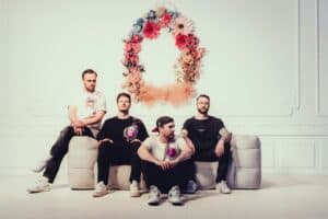 Vier Männer sitzen auf einer Couch vor einem großen Blumenkranz, symbolisieren den Kölner Karneva.