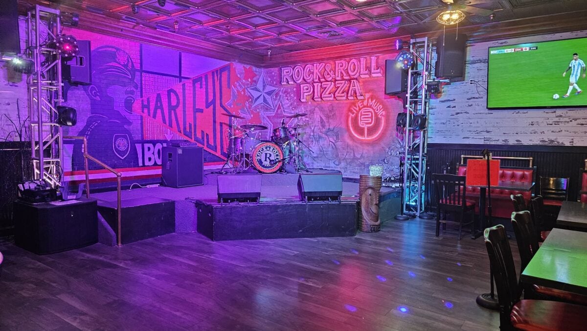 Empty stage with musical instruments at Rock & Roll Pizza. Neon signs and colorful graffiti decorate the walls. A large screen displays a sports game in the corner.